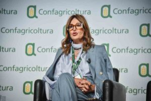 La Ministra del Turismo, Daniela Santanché, seduta ed elegantemente vestita durante un evento di Confagricoltura con tabellone con logo Confagricoltura sullo sfondo.